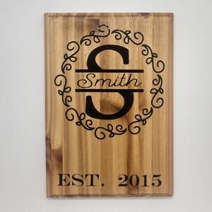 May include: A wooden sign with a black engraved initial "S" in a decorative circle with the last name "Smith" and the text "EST. 2015".