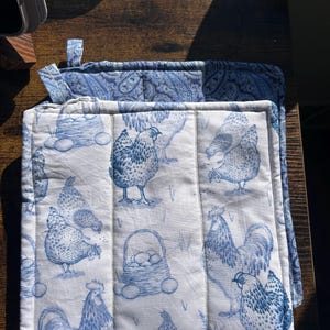 May include: A square quilted potholder featuring a blue and white toile-style print of chickens, roosters, and baskets of eggs. The fabric has a repeating pattern of poultry and farm-themed imagery, with a small fabric loop attached to the top corner.