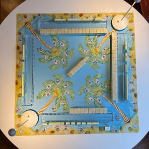 May include: A square board game with a tropical theme. The center features a light blue background with palm trees and coconuts. The board has clear plastic dividers and wooden game pieces. The edges are decorated with a beach scene.