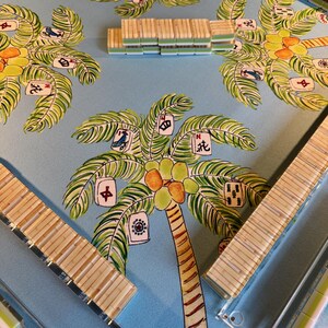 May include: A light blue Mahjong game board with a palm tree design. The board features a repeating pattern of palm trees with green fronds, coconuts, and game tiles. Several stacks of tiles and bamboo tile racks are visible.
