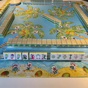 May include: A complete Mahjong set with a tropical theme. The game board features a blue background with palm tree illustrations. The tiles are white with various symbols and numbers. The set includes racks and a central spinner.