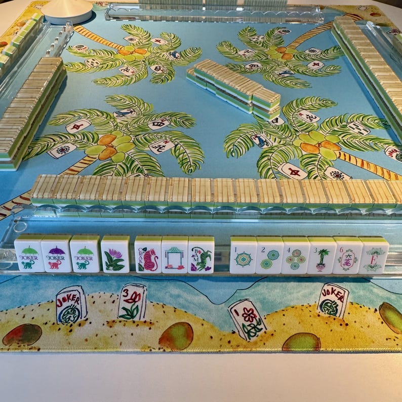 May include: A complete Mahjong set with a tropical theme. The game board features a blue background with palm tree illustrations. The tiles are white with various symbols and numbers. The set includes racks and a central spinner.