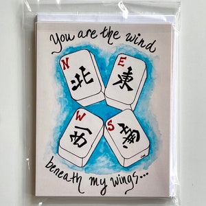 May include: A white card with a blue watercolor background and four white mahjong tiles with black and red Chinese characters. The tiles are arranged in a cross shape with the characters facing outward. The text "You are the wind beneath my wings..." is written in black cursive below the tiles.