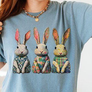 Puede incluir: Una camiseta azul claro con un gráfico de tres conejos sentados uno al lado del otro. Cada conejo tiene un patrón único en su cuerpo, incluyendo cuadros azules y blancos, flores y diseños de tartán multicolor, con orejas de tono rosado.