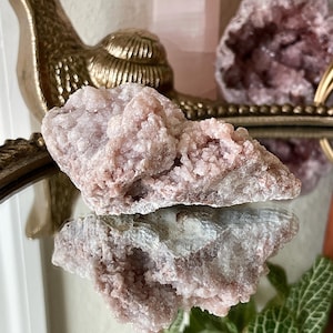 May include: A light pink, crystalline mineral specimen with a rough, textured surface. The mineral is reflected in a mirrored surface, with a gold-colored decorative element in the background. The overall aesthetic is natural and earthy.
