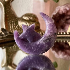 May include: A carved purple crystal moon and star figurine. The crescent moon shape cradles a five-pointed star. The crystal has a polished surface and is displayed on a reflective surface.