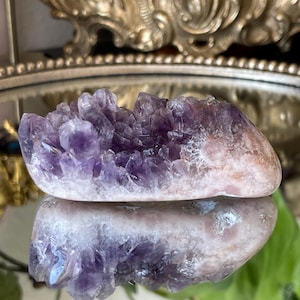 May include: A polished amethyst crystal cluster, showcasing deep purple and pale lavender hues. The crystal formation is set against a reflective surface, highlighting its natural beauty and intricate structure. A decorative item for home decor.