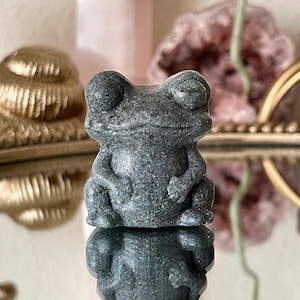 May include: A detailed, gray stone frog figurine with large, expressive eyes and a textured surface. The frog is in a seated position, with its arms and legs folded. The figurine is reflected in a mirrored surface, creating a symmetrical image.