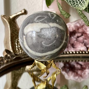 May include: A grey and white patterned sphere sits on a gold-coloured butterfly-shaped stand. The sphere has a polished surface with swirling patterns. The stand is made of metal and has a reflective surface.