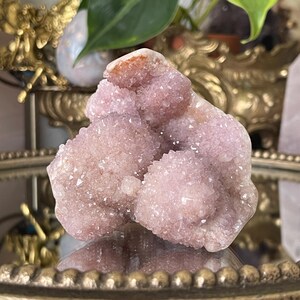 May include: A cluster of pink amethyst crystals with a rough, natural texture. The crystals are arranged in a cluster, with some of the crystals pointing upwards. The crystals are a light pink color, with some areas of darker pink. The crystals are translucent, and the light shines through them.