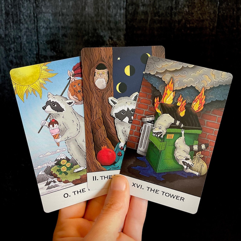 Op de afbeelding: Drie tarotkaarten met illustraties van wasberen. De eerste kaart is gelabeld "0. De Dwaas" en toont een wasbeer die een ijsje vasthoudt. De tweede kaart is gelabeld "II. De Hogepriesteres" en toont een wasbeer die voor een boom staat met een uil in een nest. De derde kaart is gelabeld "XVI. De Toren" en toont een wasbeer die voor een brandend gebouw staat.