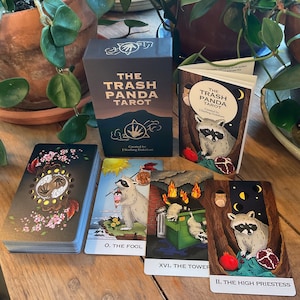 Op de afbeelding: Een zwarte doos met gouden letters met de tekst "The Trash Panda Tarot" en een kleinere doos met dezelfde tekst. Er liggen drie tarotkaarten op een houten tafel, &eacute;&eacute;n met een wasbeer die een beker ijs vasthoudt, een andere met een wasbeer in een vuilnisbak en de laatste met een wasbeer die een granaatappel vasthoudt.