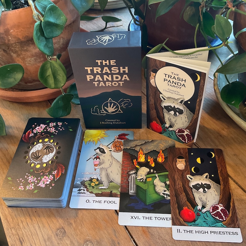 Op de afbeelding: Een zwarte doos met gouden letters met de tekst "The Trash Panda Tarot" en een kleinere doos met dezelfde tekst. Er liggen drie tarotkaarten op een houten tafel, &eacute;&eacute;n met een wasbeer die een beker ijs vasthoudt, een andere met een wasbeer in een vuilnisbak en de laatste met een wasbeer die een granaatappel vasthoudt.