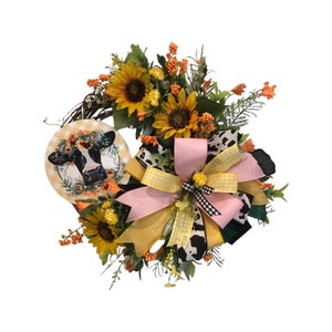 May include: A farmhouse-style wreath with a black and white cow face in the center. The wreath is decorated with sunflowers, yellow and pink gingham ribbon, and a black and white cow print ribbon.
