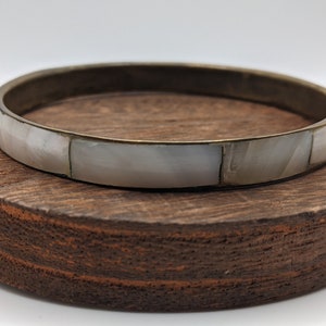 May include: A brass bangle bracelet with a white mother of pearl inlay. The bracelet is on a wooden surface.