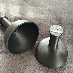 Può includere: Due oggetti neri a forma di campana con uno stelo. Uno è aperto, rivelando l'interno. L'altro ha il testo "2.90 Inches" sulla parte superiore. Gli oggetti sono realizzati in un materiale strutturato e poggiano su una superficie grigia.