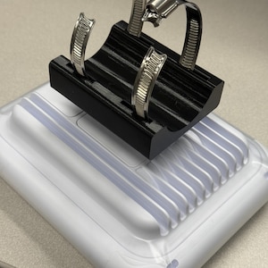 May include: Two silver-colored metal worm gear hose clamps with slotted bands and screw-tightening mechanisms, positioned upright in a black plastic holder that is mounted on a white rectangular base with a ribbed, translucent top surface.