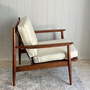 May include: Mid-century modern armchair with a wooden frame and cream-colored cushions. The chair features a low profile, angled legs, and armrests. The cushions have a green trim. The chair is placed on a patterned rug.