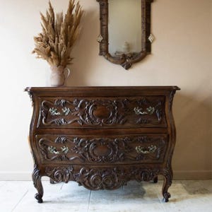 May include: A dark brown, ornately carved wooden chest of drawers with two drawers. The chest of drawers has a curved front and sits on four legs. It is in front of a beige wall with a decorative mirror above it. A dried flower arrangement is in a vase to the left of the chest of drawers.
