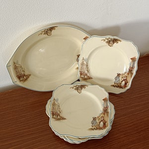 May include: Set of four vintage ceramic plates with a cream background and a blue rim. The plates feature a brown and white illustration of a town scene with people walking down the street.