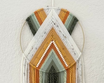 May include: A macrame wall hanging with a circular wooden frame and a geometric pattern of white, orange, and green yarn. The hanging has long strands of yarn in the same colors.