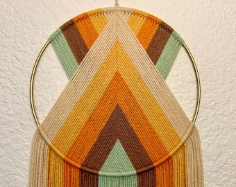 May include: A circular macrame wall hanging with a geometric pattern in shades of brown, orange, and green. The macrame is attached to a gold metal hoop and has long fringe strands.