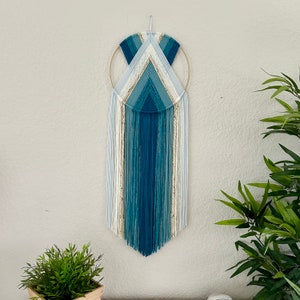 May include: A blue, white, and beige macrame wall hanging with a gold hoop. The macrame is in the shape of a teardrop with a geometric pattern.