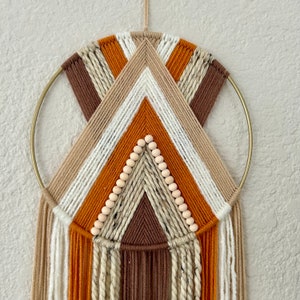 May include: A macrame wall hanging with a geometric design in shades of brown, beige, and orange. The hanging features a circular wooden hoop and a fringe of yarn.