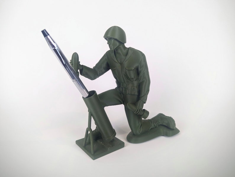 Peut inclure: Une figurine de soldat en plastique vert agenouill&eacute;e tenant un stylo dans un tube de mortier. Le tube de mortier est sur une base et le stylo sort du haut.
