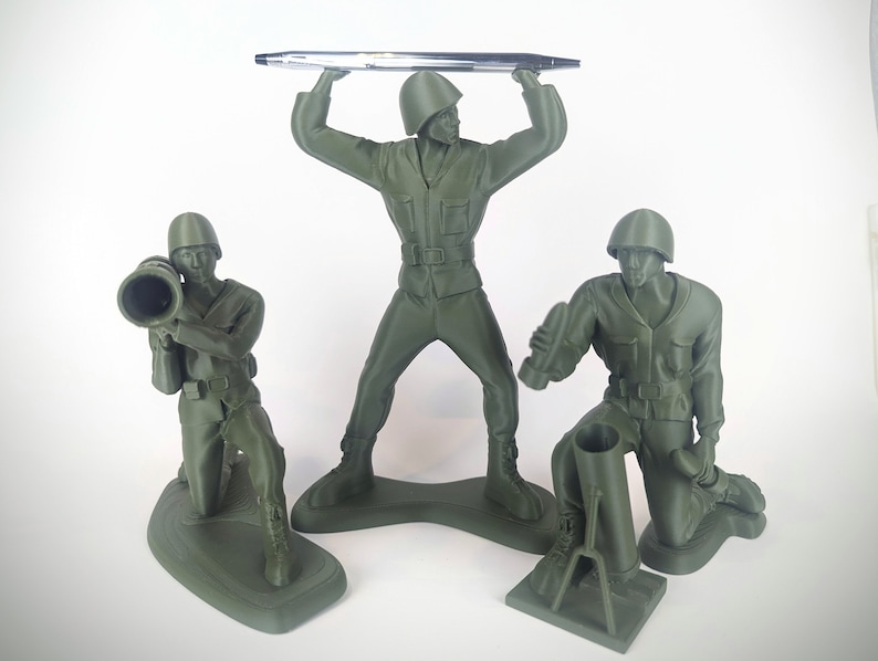 Peut inclure: Trois soldats en plastique vert, l'un tenant un stylo, l'autre un bazooka et le dernier un mortier. Les soldats sont tous dans des poses diff&eacute;rentes et se tiennent sur une surface blanche.