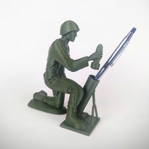 Peut inclure: Une figurine de soldat en plastique vert tenant un stylo dans un porte-stylo en forme de mortier.