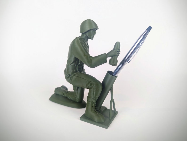 Peut inclure: Une figurine de soldat en plastique vert tenant un stylo dans un porte-stylo en forme de mortier.