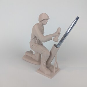 Peut inclure: Une figurine en plastique beige d'un soldat agenouill&eacute; tenant un stylo dans un tube de mortier. Le soldat porte un casque et une ceinture de munitions. Le tube de mortier est sur un tr&eacute;pied.