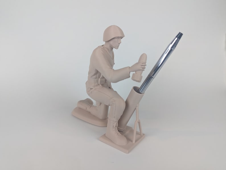 Peut inclure: Une figurine en plastique beige d'un soldat agenouill&eacute; tenant un stylo dans un tube de mortier. Le soldat porte un casque et une ceinture de munitions. Le tube de mortier est sur un tr&eacute;pied.
