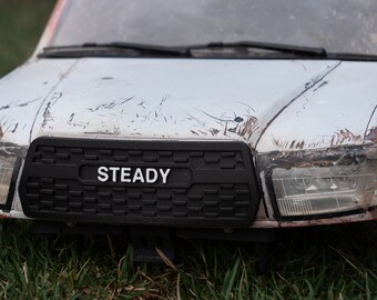 May include: Close-up of a white toy truck with a black grill and the word "STEADY" printed on it. The truck has a weathered, worn look, suggesting it has been used for off-road driving.
