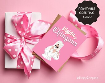 May include: A pink greeting card with the text "Viejita y Chingona" and an illustration of a woman in a bathrobe and sunglasses. The card is next to a wrapped gift with a pink polka dot ribbon.