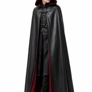 May include: A floor-length black hooded cloak with a deep red interior lining. The garment features a front buckle closure at the neck and a structured collar, worn over a matching black long-sleeved tunic and black trousers with black boots.