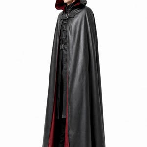 May include: A floor-length black hooded cloak featuring a deep red interior lining. The garment includes a front buckle closure at the neck and a series of vertical decorative toggle fasteners down the center, worn over black trousers and boots.