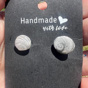 May include: A pair of stud earrings featuring small, circular, natural grey and white spiral seashell pieces attached to a black rectangular display card with the white printed text "Handmade with love" and a small white heart icon.