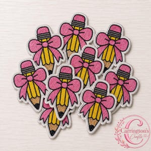 Könnte beinhalten: Eine Sammlung von gestickten Aufnähern in Form von gelben Bleistiften mit rosa Schleifen. Jeder Bleistift hat einen grauen Radiergummi, einen gelben Körper und eine dunkelbraune Spitze. Die rosa Schleifen sitzen oben am Bleistift. Das Logo Carrington Crafts ist sichtbar.