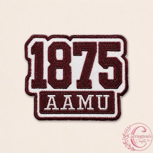 Op de afbeelding: Een rechthoekige geborduurde patch met de bordeauxrode cijfers 1875 boven de bordeauxrode tekst AAMU, alles omlijnd in wit en bordeauxrood. Het ontwerp heeft een dikke rand en een textuur van stof met duidelijke, vetgedrukte letters en cijfers.