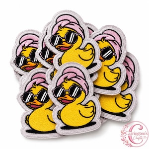Peut inclure: Une collection d'écussons brodés représentant un canard en caoutchouc jaune portant des lunettes de soleil noires et une serviette rose enroulée autour de la tête. Chaque écusson a une bordure gris clair et un motif cousu détaillé.