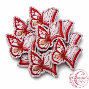 May include: A collection of embroidered patches featuring a butterfly design where the wings are shaped like open books. The patches are colored in red, white, and gold thread with a white border. The logo Carrington's Crafts is in the corner.