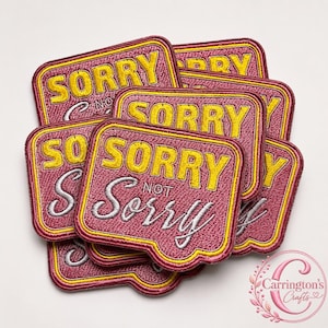 Può includere: Una collezione di toppe ricamate rettangolari con angoli arrotondati. Ogni toppa presenta uno sfondo rosa scuro con un bordo giallo e il testo SORRY in lettere maiuscole gialle sopra la parola NOT e Sorry in corsivo color argento.