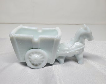 May include: White milk glass horse-drawn carriage figurine. The carriage has a single wheel and a rectangular bed. The horse is standing with its head turned to the side.