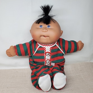 May include: A Cabbage Patch Kid doll wearing a red and green striped sleeper with a black mohawk.
