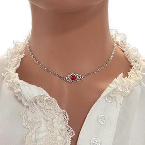 May include: Silver necklace with a central oval red gemstone, surrounded by a floral motif. The chain has a twisted rope design. The necklace is displayed against a white blouse with lace trim.