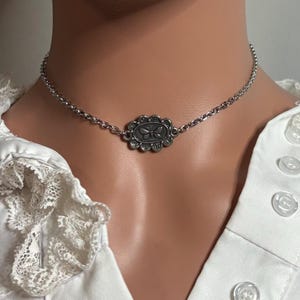 Puede incluir: Un collar de cadena en tono plateado que presenta un colgante central de forma ovalada con un borde festoneado y un motivo de mariposa grabado en el centro. El collar descansa sobre el cuello, mostrando un diseño de cadena de eslabones finos.