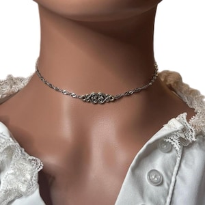 May include: A silver-toned choker necklace featuring a delicate twisted chain and a central decorative metal component with an intricate, symmetrical scrollwork pattern. The piece rests against the neck, showcasing a polished metallic finish.