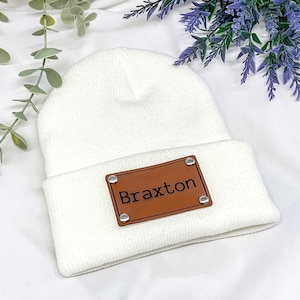 May include: A white knit beanie featuring a rectangular brown faux leather patch centered on the cuff. The patch is secured with four silver-toned metal rivets at the corners and has the name Braxton embossed in black lettering on the front.